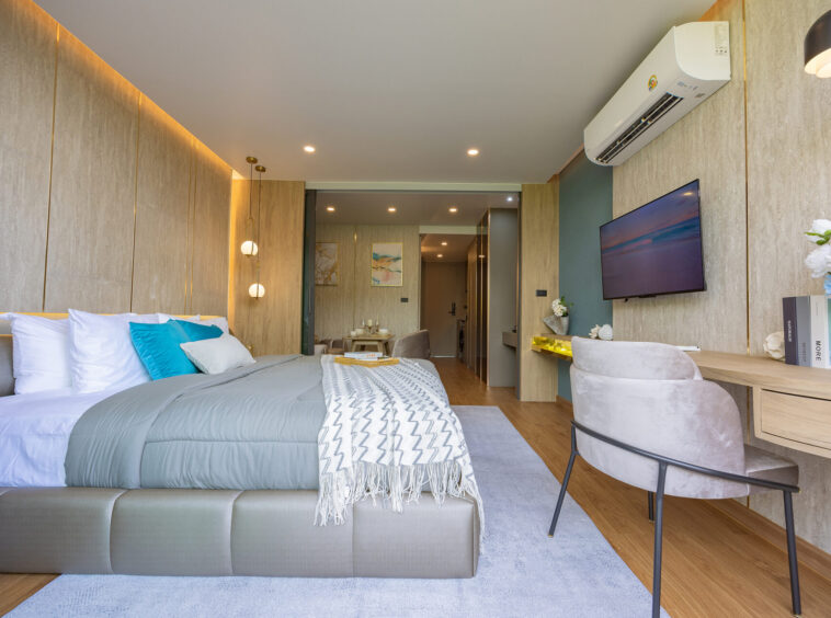 Modern hotel bedroom with a large bed, grey upholstery, and blue-green pillows facing a wall-mounted TV across the room.