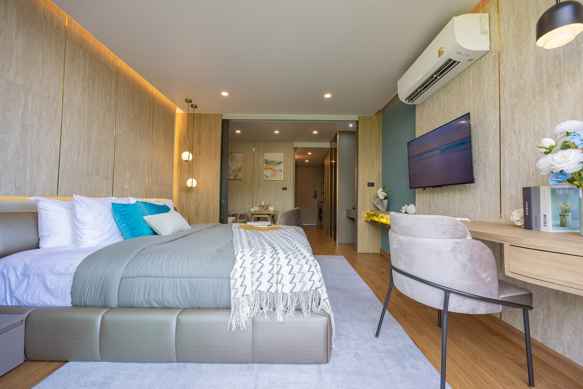 Modern hotel bedroom with a large bed, grey upholstery, and blue-green pillows facing a wall-mounted TV across the room.