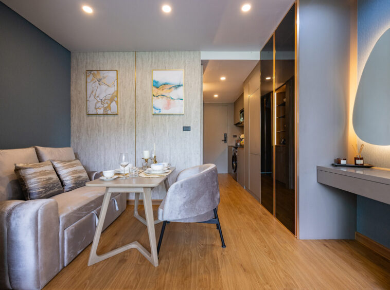 Open-plan living area with a beige sofa, decorative cushions, and a light wood dining table set for four in a contemporary apartment.