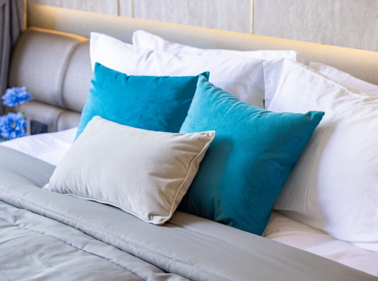 A neatly made bed with white sheets and blue and beige throw pillows under warm ambient lighting.
