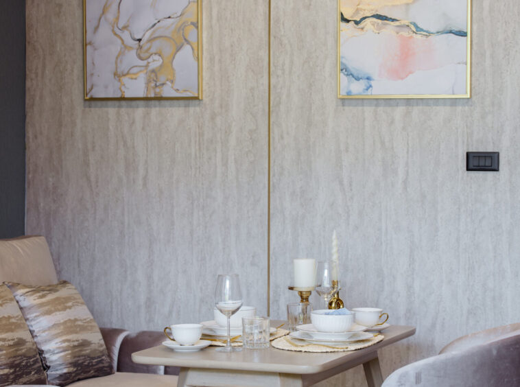 Two gold-framed abstract paintings on a light textured wall above a small dining table set with white dishes, glasses, and candles.