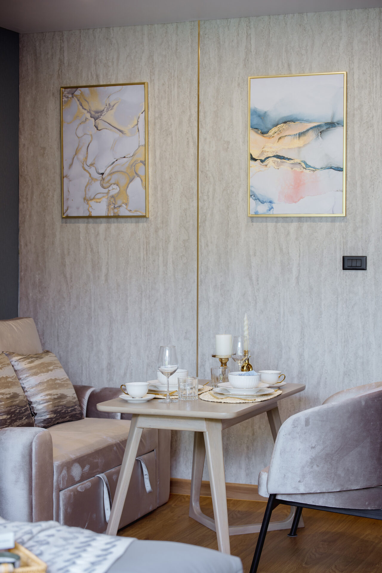 Two gold-framed abstract paintings on a light textured wall above a small dining table set with white dishes, glasses, and candles.