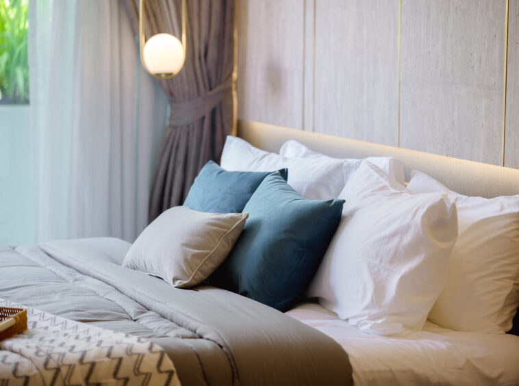 Neatly made bed with white linens and blue and beige pillows in a modern hotel room, with a soft-lit headboard and curtains nearby.