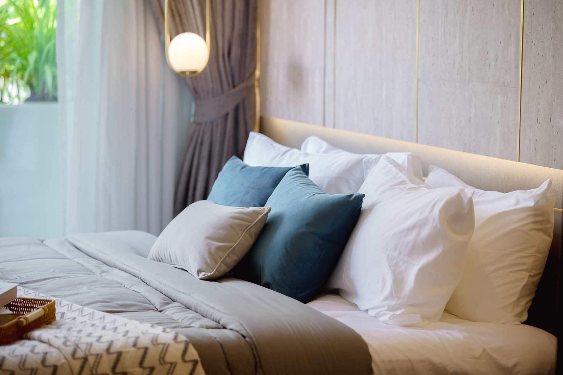 Neatly made bed with white linens and blue and beige pillows in a modern hotel room, with a soft-lit headboard and curtains nearby.