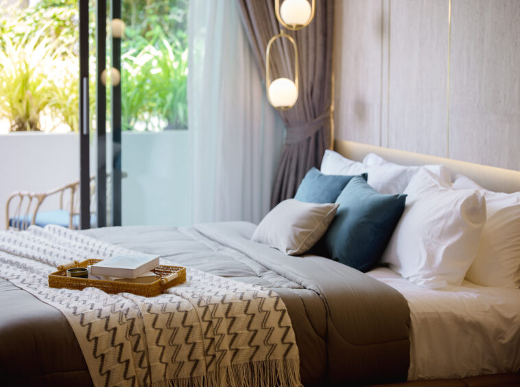 Luxurious hotel bedroom with a large bed, beige bedding, and blue-green pillows, a woven tray and book at the foot of the bed; glass doors reveal greenery outside.