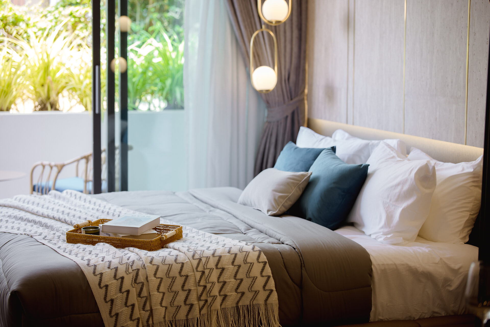 Luxurious hotel bedroom with a large bed, beige bedding, and blue-green pillows, a woven tray and book at the foot of the bed; glass doors reveal greenery outside.