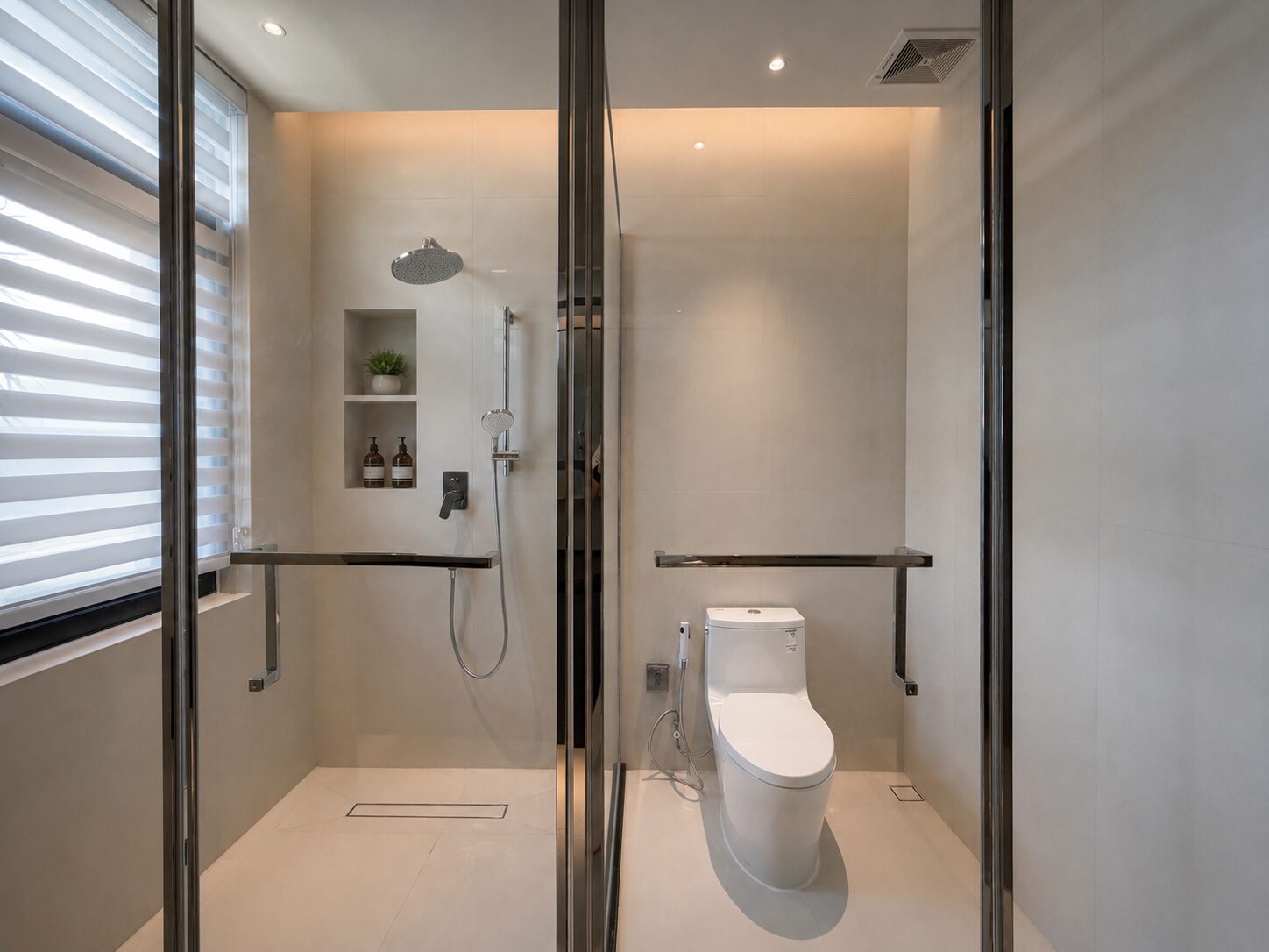 Modern accessible bathroom with a walk-in shower, toilet, grab bars, and a glass partition; neutral-toned tiles and a window with blinds.