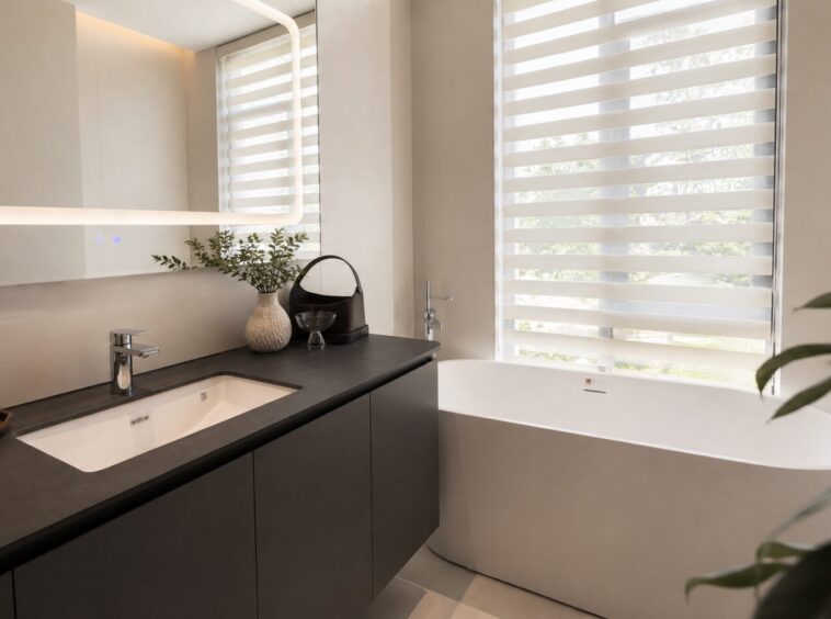 Modern bathroom with dark vanity, white undermount sink, chrome faucet, and decorative items on countertop beside a freestanding white tub by large striped blinds.