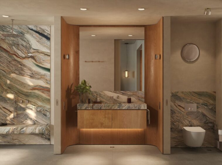 Spa-style bathroom featuring marble walls, a wooden vanity, and a wall-hung toilet with a large mirror above the sink; natural light from the left windowed wall.