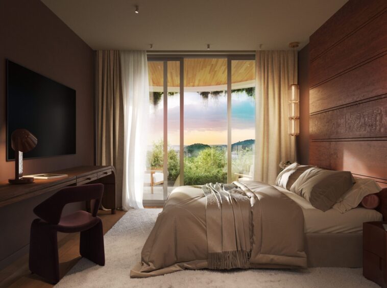 Bedroom with a large bed in beige bedding, beige curtains, and glass doors opening to a balcony with a sunset over greenery.