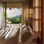 Hotel bedroom with beige bedding, copper ribbed column lamps, and a view of lush greenery and distant mountains through glass doors.