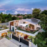 Aerial view of a modern multi-level house with a solar-panel roof, spacious patio, and surrounding tropical trees and neighborhood.”