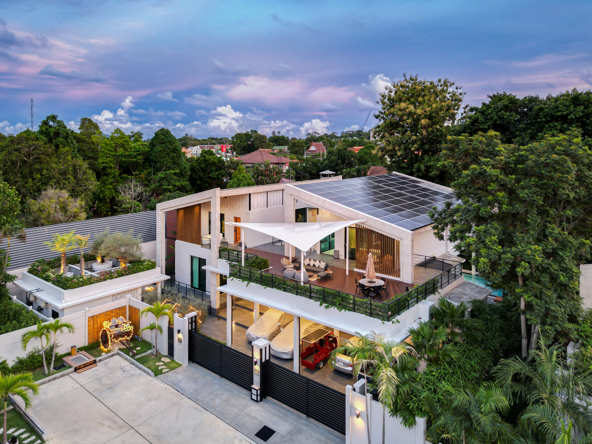 Aerial view of a modern multi-level house with a solar-panel roof, spacious patio, and surrounding tropical trees and neighborhood.”