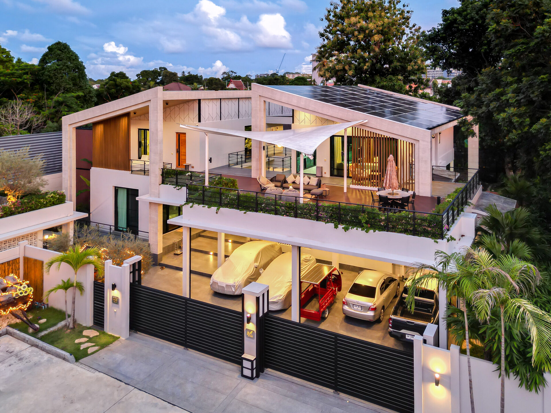 Modern villa with rooftop terrace, glass walls, and solar panels; multiple parked cars on a gated, multi-level property.