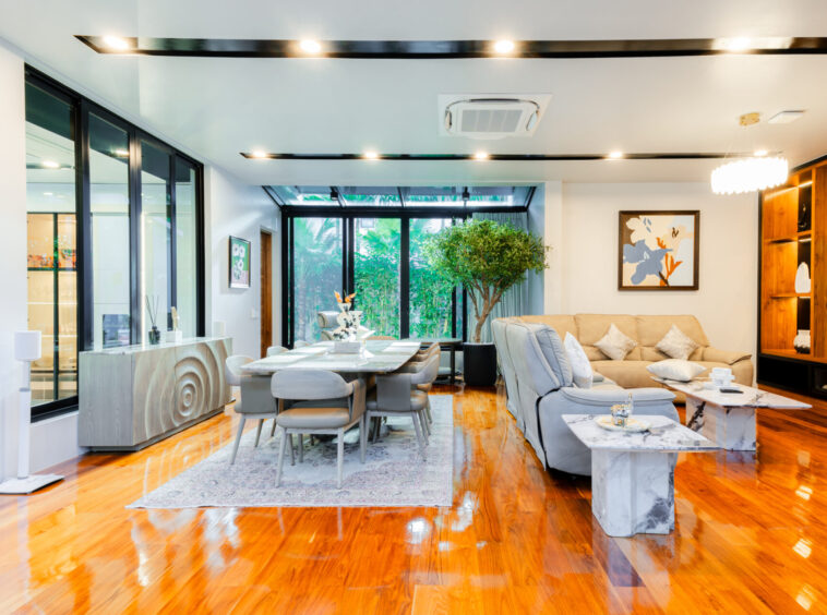 Open-plan living and dining room with glass walls, warm hardwood floors, a gray dining table, and a beige sofa framed by greenery and art.