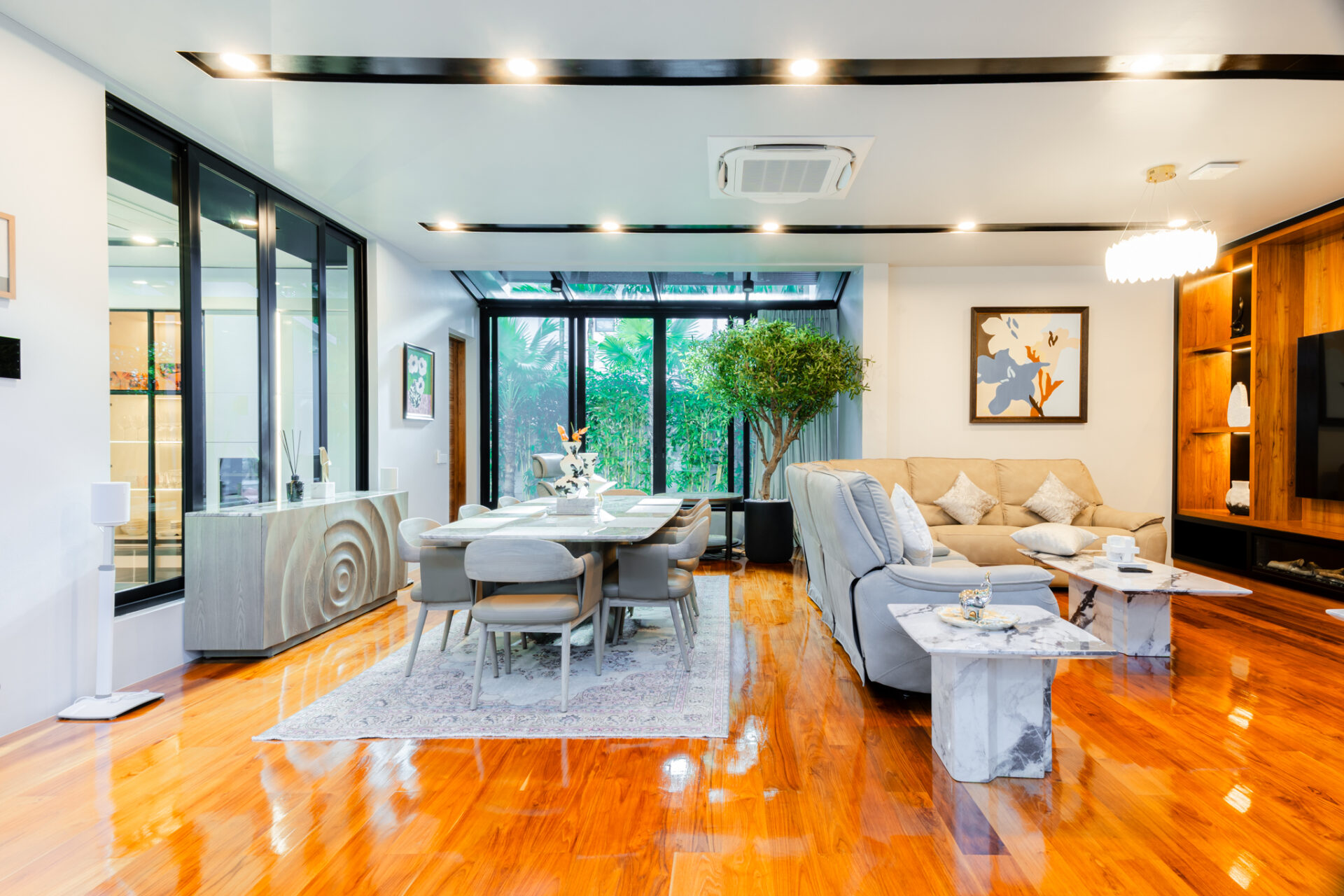 Open-plan living and dining room with glass walls, warm hardwood floors, a gray dining table, and a beige sofa framed by greenery and art.