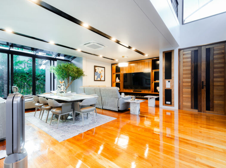 Open-plan living and dining area with glass walls, a large dining table, and a gray sofa under bright overhead lighting.