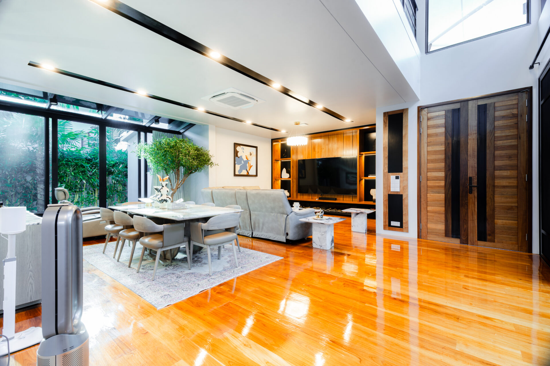 Open-plan living and dining area with glass walls, a large dining table, and a gray sofa under bright overhead lighting.