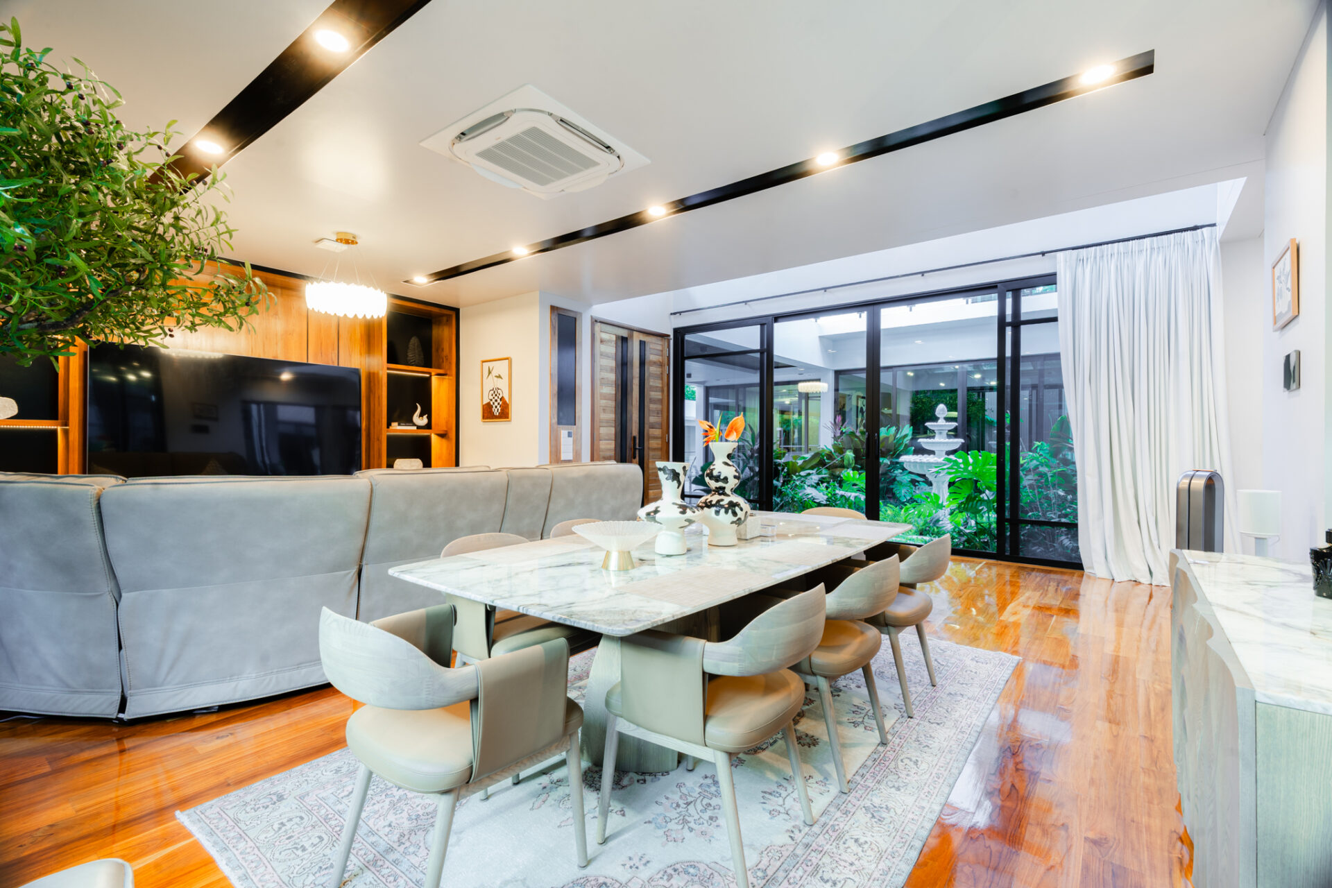 Open-plan living and dining area with a marble table, gray sofa, and large glass doors opening to a garden.