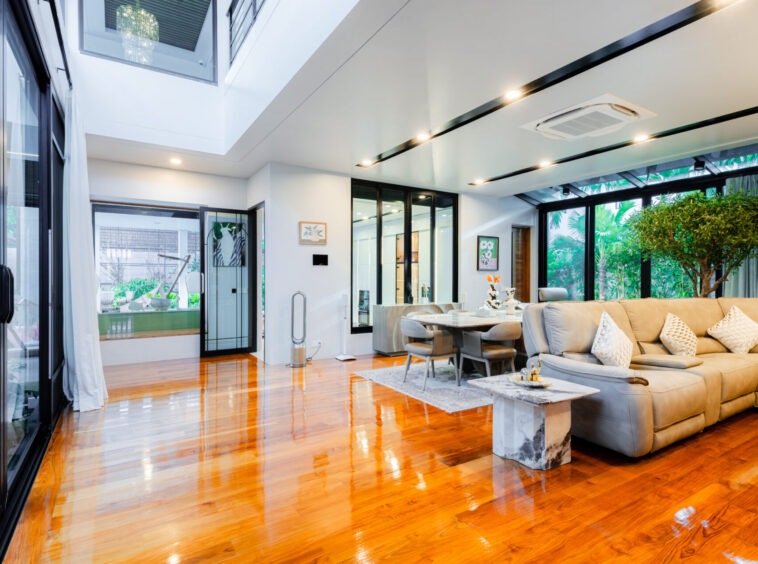 Bright open-concept living room with glass walls, warm wood floor, beige sectional, and indoor plants.