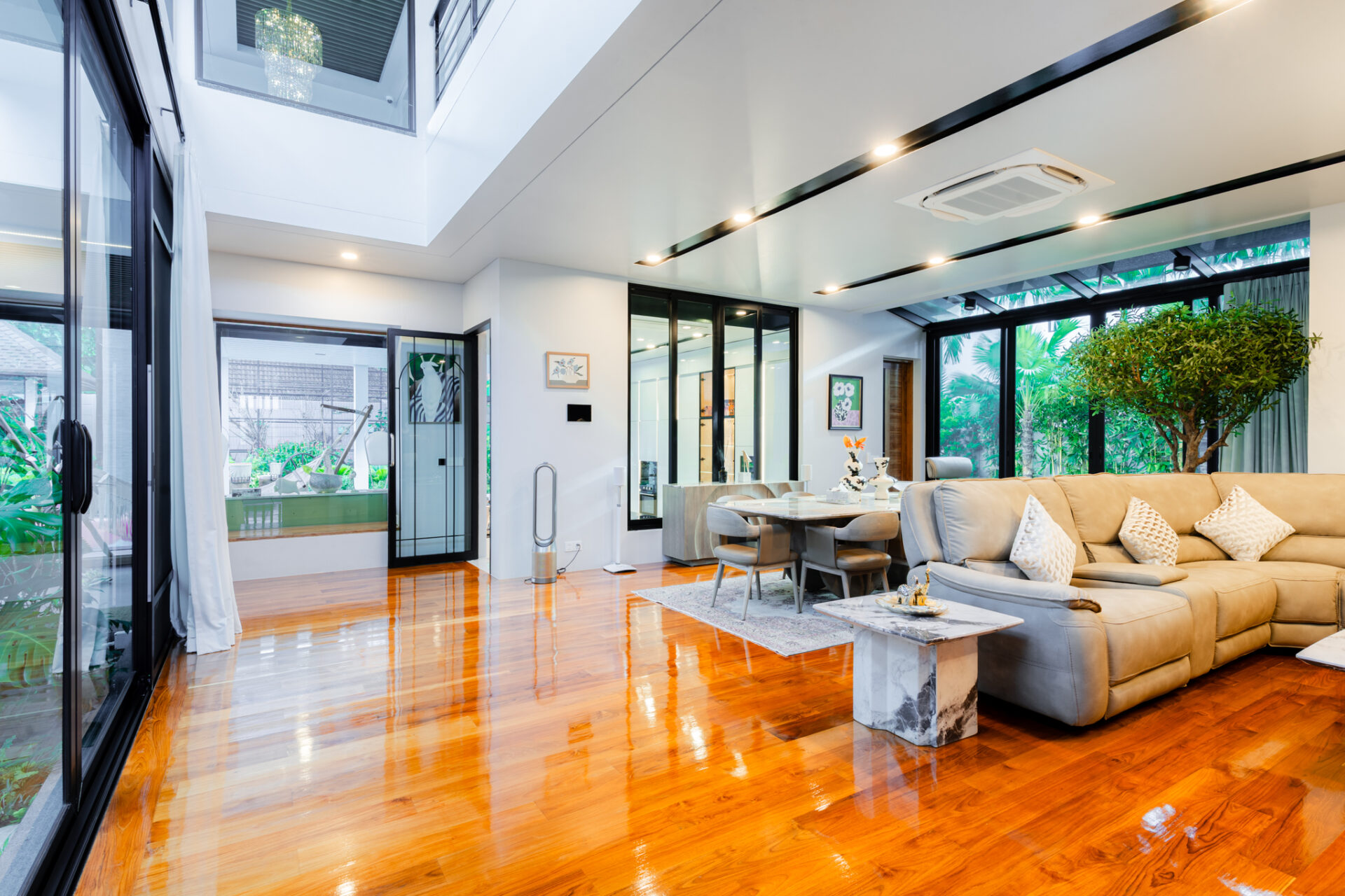 Bright open-concept living room with glass walls, warm wood floor, beige sectional, and indoor plants.