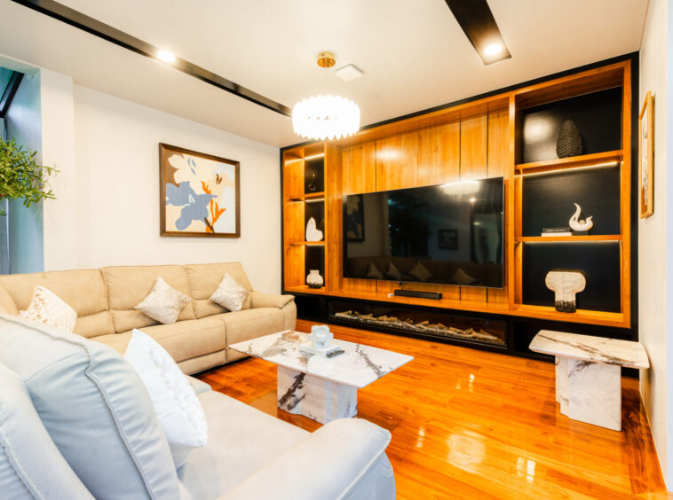 Bright living room with a beige sectional sofa, marble coffee tables, and a large flat-screen TV set into a warm wood entertainment wall.