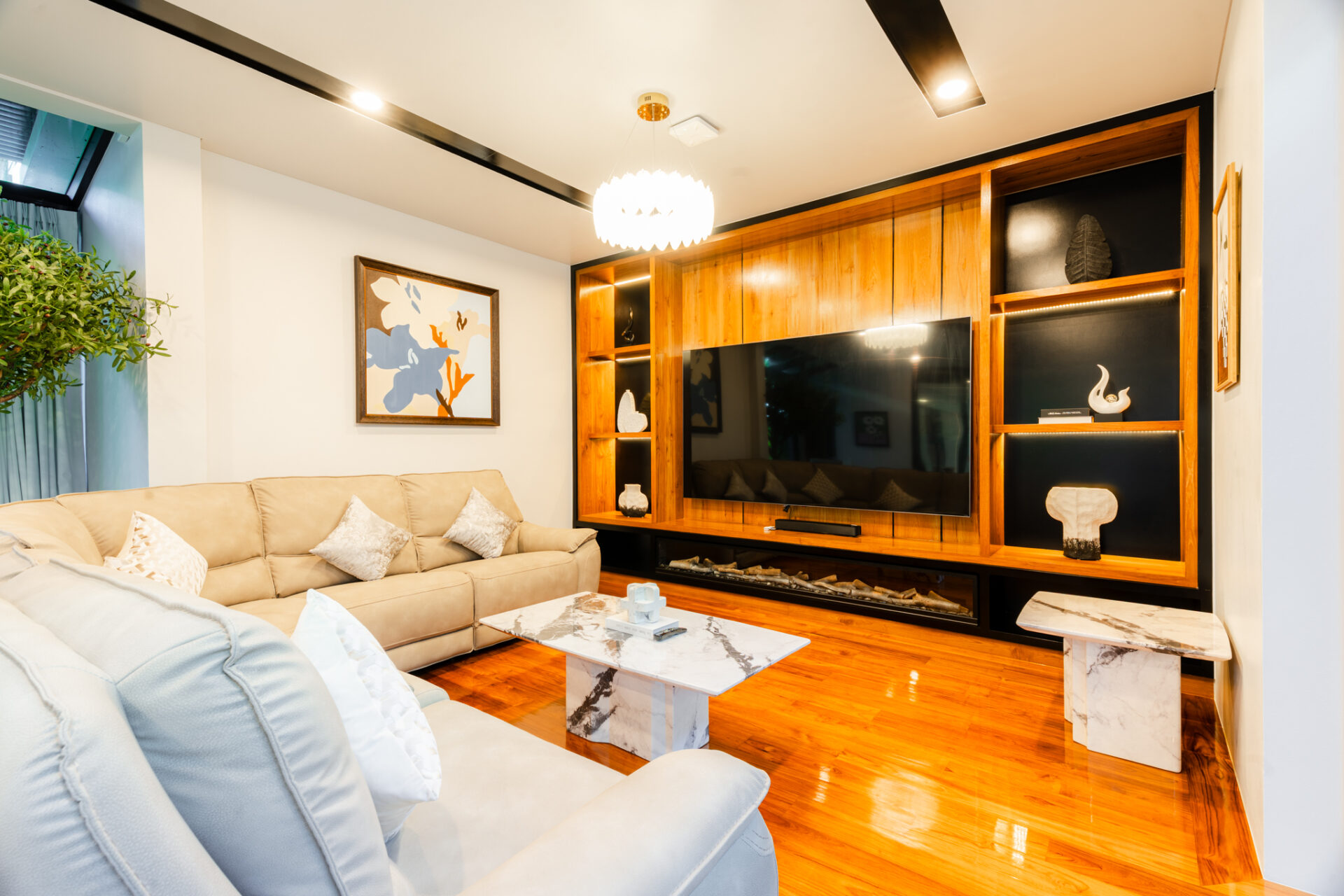 Bright living room with a beige sectional sofa, marble coffee tables, and a large flat-screen TV set into a warm wood entertainment wall.