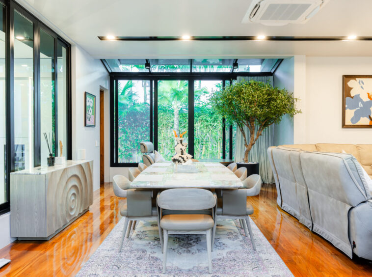 Modern dining room with a long marble table, gray chairs, and a large indoor tree next to a beige sofa; glass wall reveals a tropical garden outside.
