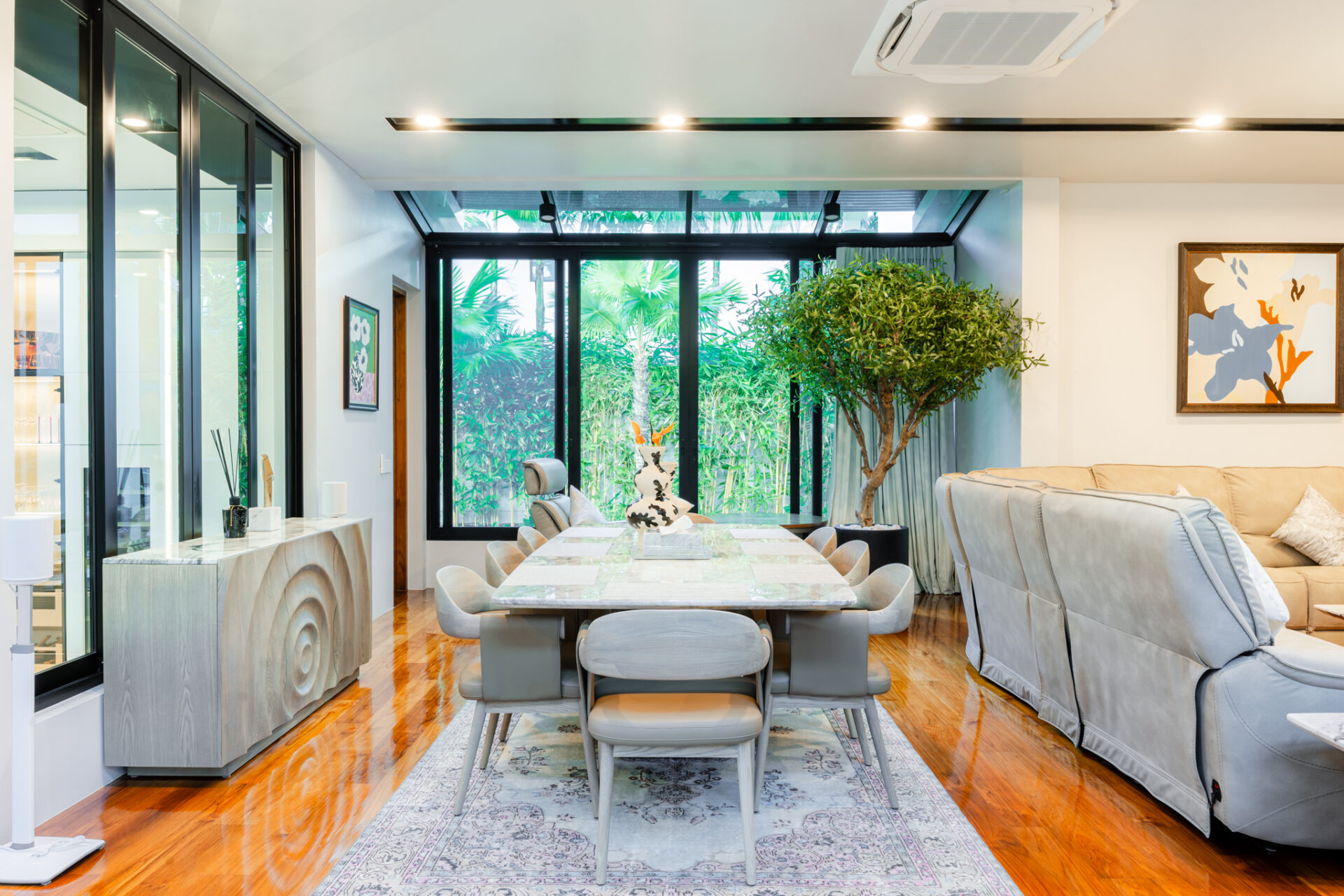 Modern dining room with a long marble table, gray chairs, and a large indoor tree next to a beige sofa; glass wall reveals a tropical garden outside.