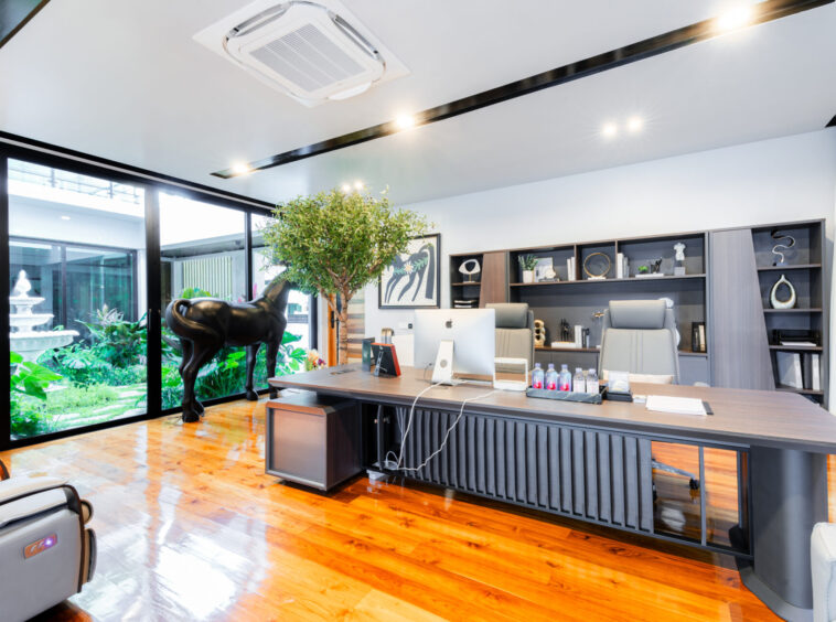 Reception area of a modern office with a long dark wood desk, computer, and built-in shelves; a black horse sculpture and greenery nearby under bright overhead lighting visible through large windows.