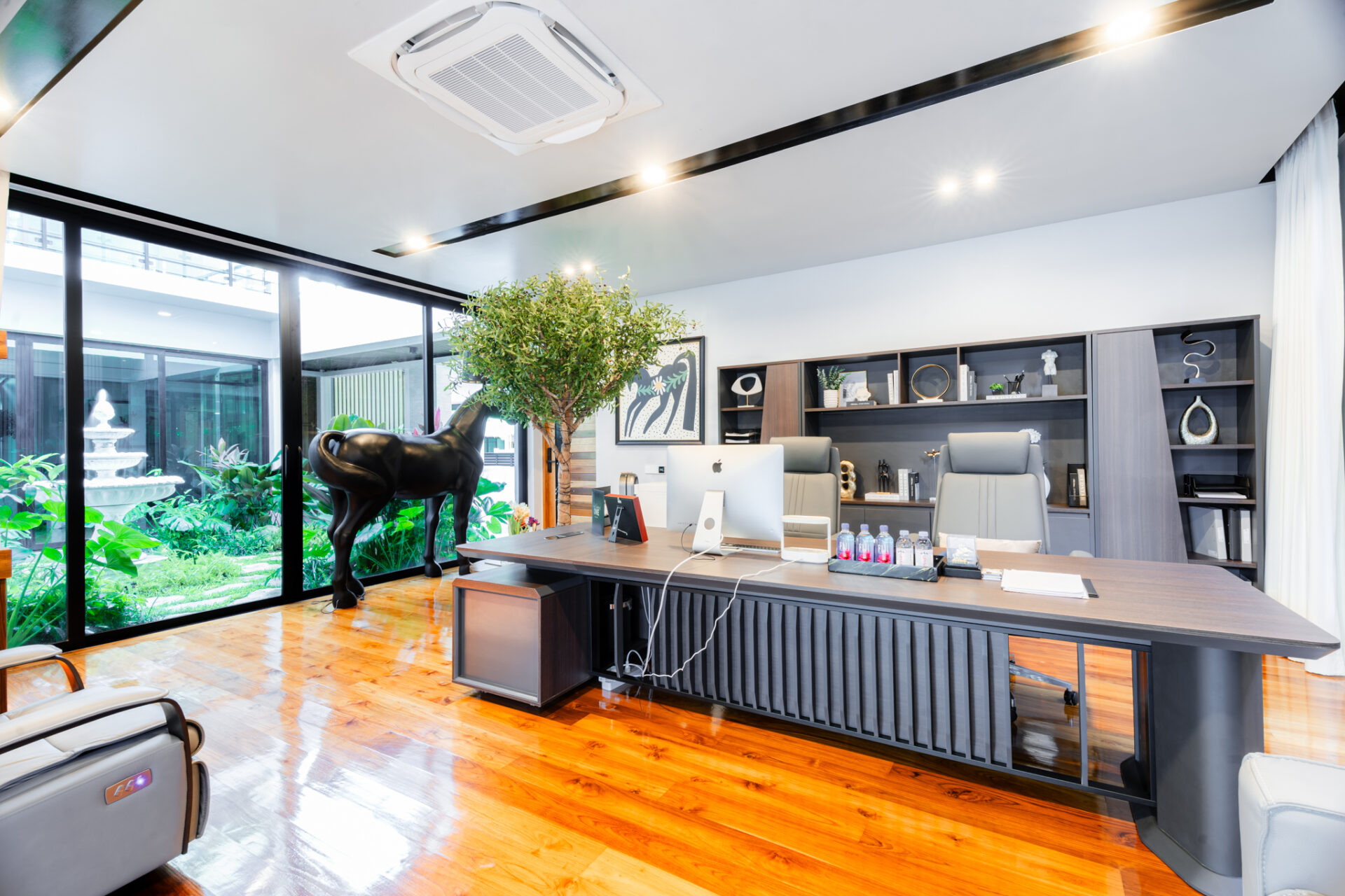 Reception area of a modern office with a long dark wood desk, computer, and built-in shelves; a black horse sculpture and greenery nearby under bright overhead lighting visible through large windows.