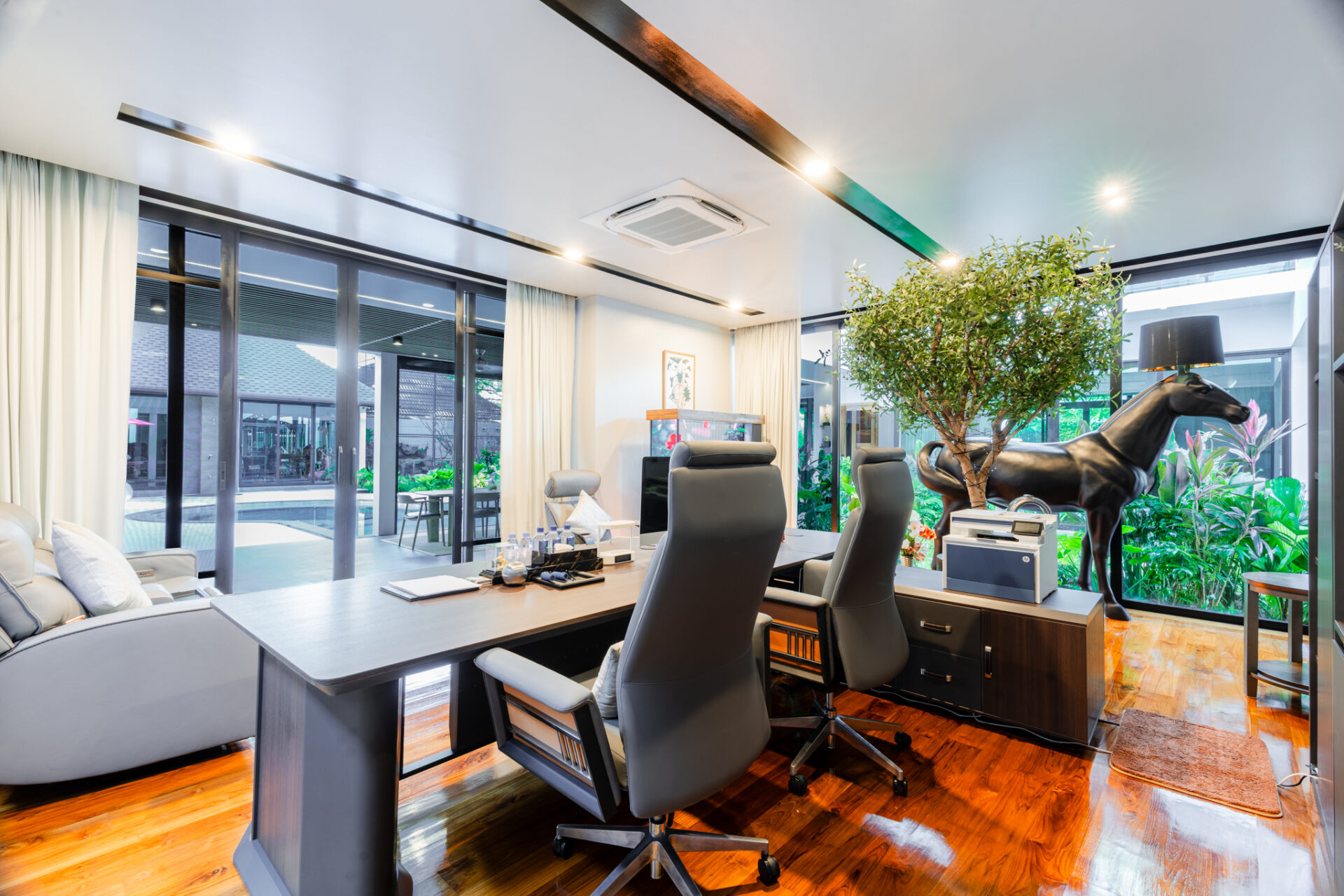 Modern office with a long desk, black ergonomic chairs, and a life-size black horse statue among lush plants by large glass walls.