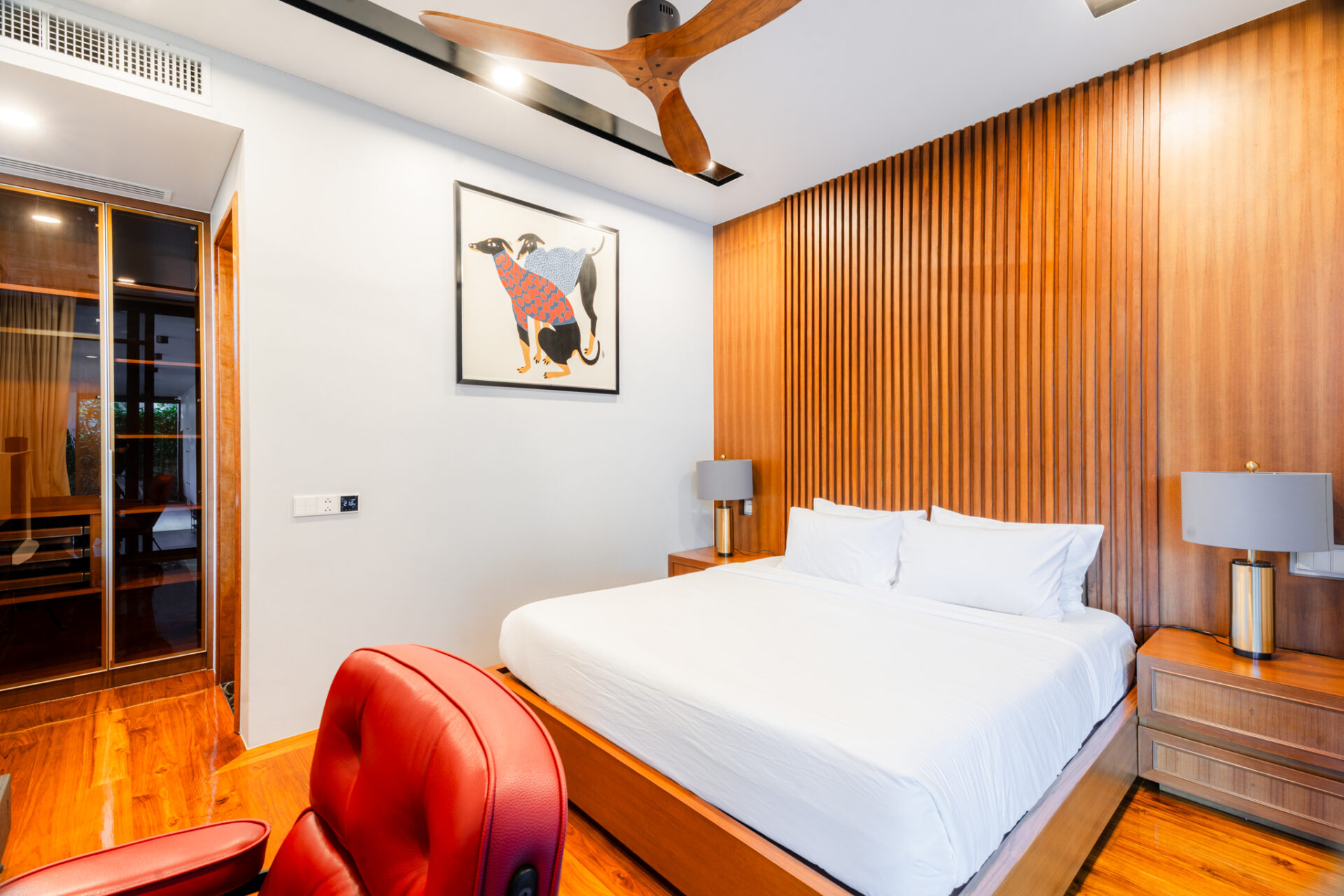 Bedroom with white bedding, wood slat accent wall, matching nightstands and lamps; red leather chair in foreground.