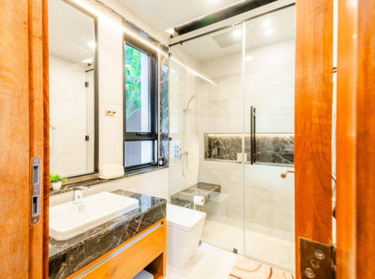 Modern contemporary bathroom with a glass shower enclosure, double-sink vanity, and dark marble countertops accented by warm wood cabinetry and a small plant near the sink window.
