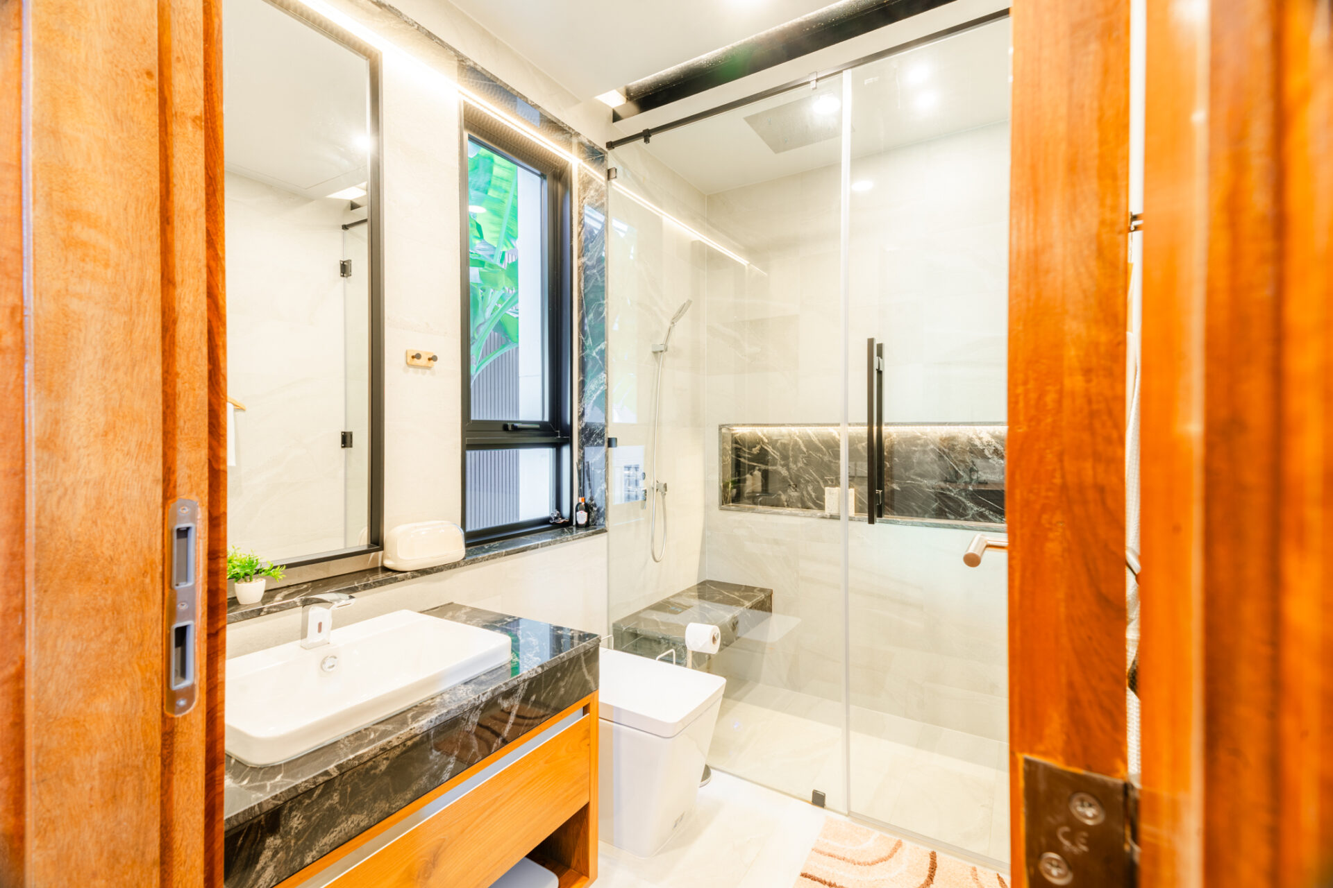 Modern contemporary bathroom with a glass shower enclosure, double-sink vanity, and dark marble countertops accented by warm wood cabinetry and a small plant near the sink window.