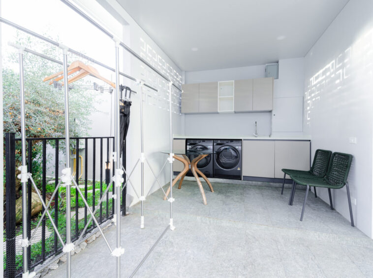 Modern indoor laundry room with two front-loading washers, grey cabinets, and a metal clothes-drying rack; garden visible outside through a railing.