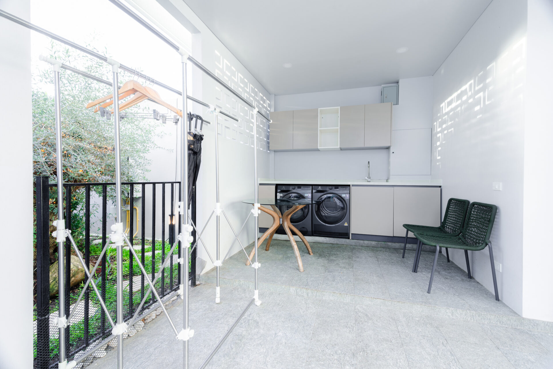 Modern indoor laundry room with two front-loading washers, grey cabinets, and a metal clothes-drying rack; garden visible outside through a railing.