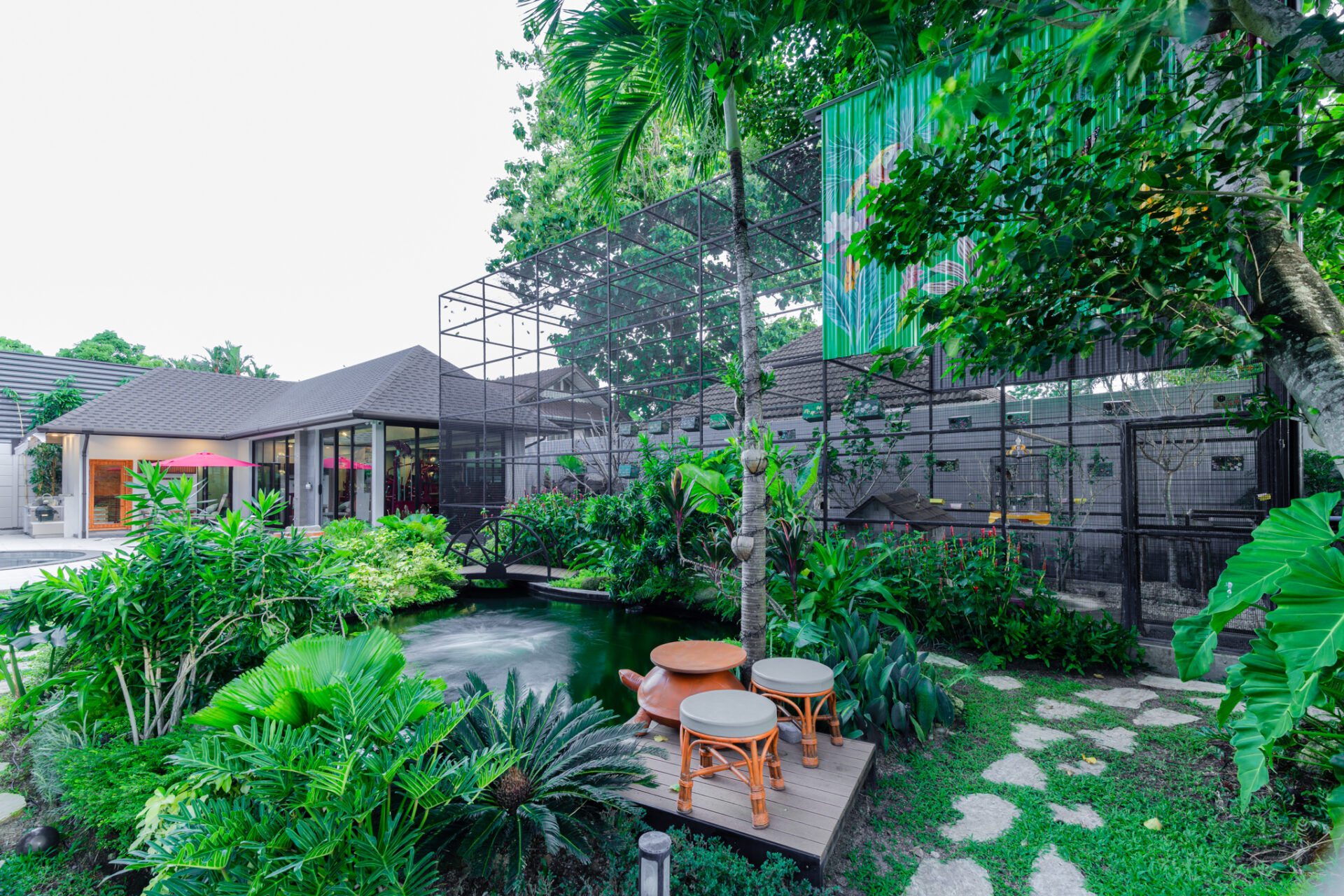 Lush tropical garden with a wooden deck and orange-stemmed stools beside a small pond, with a modern house in the background.