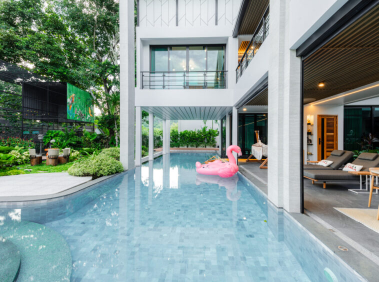 Modern white building with glass walls surrounding a rectangular pool; a pink flamingo float on the water near the outdoor lounge area.