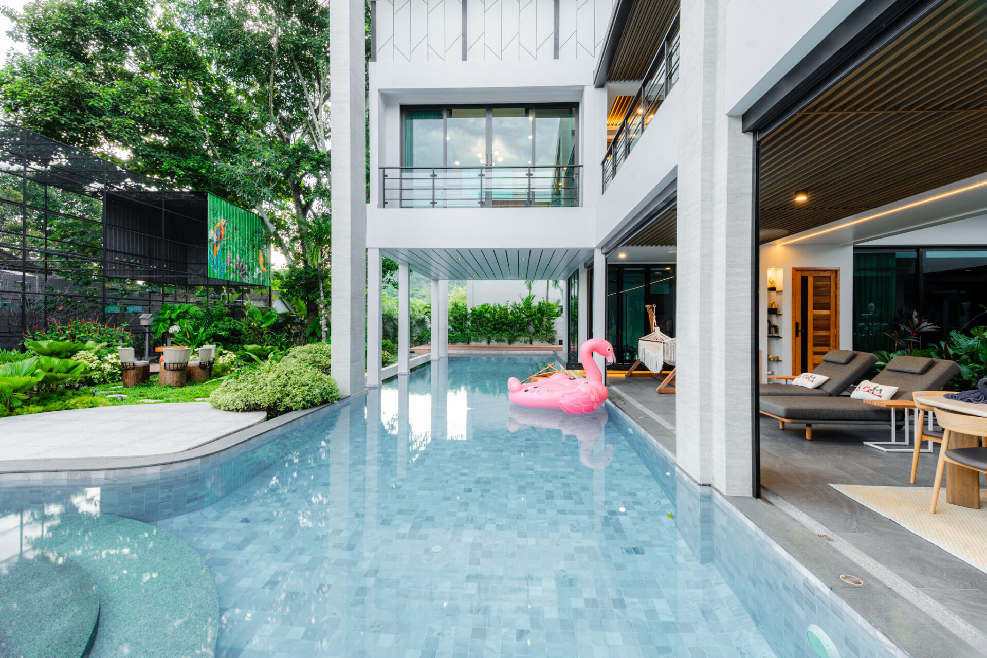 Modern white building with glass walls surrounding a rectangular pool; a pink flamingo float on the water near the outdoor lounge area.