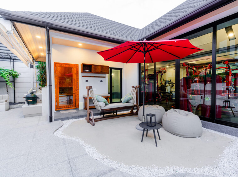 Outdoor patio with a red umbrella, wooden seating, and bean bags beside a glass-walled gym area.