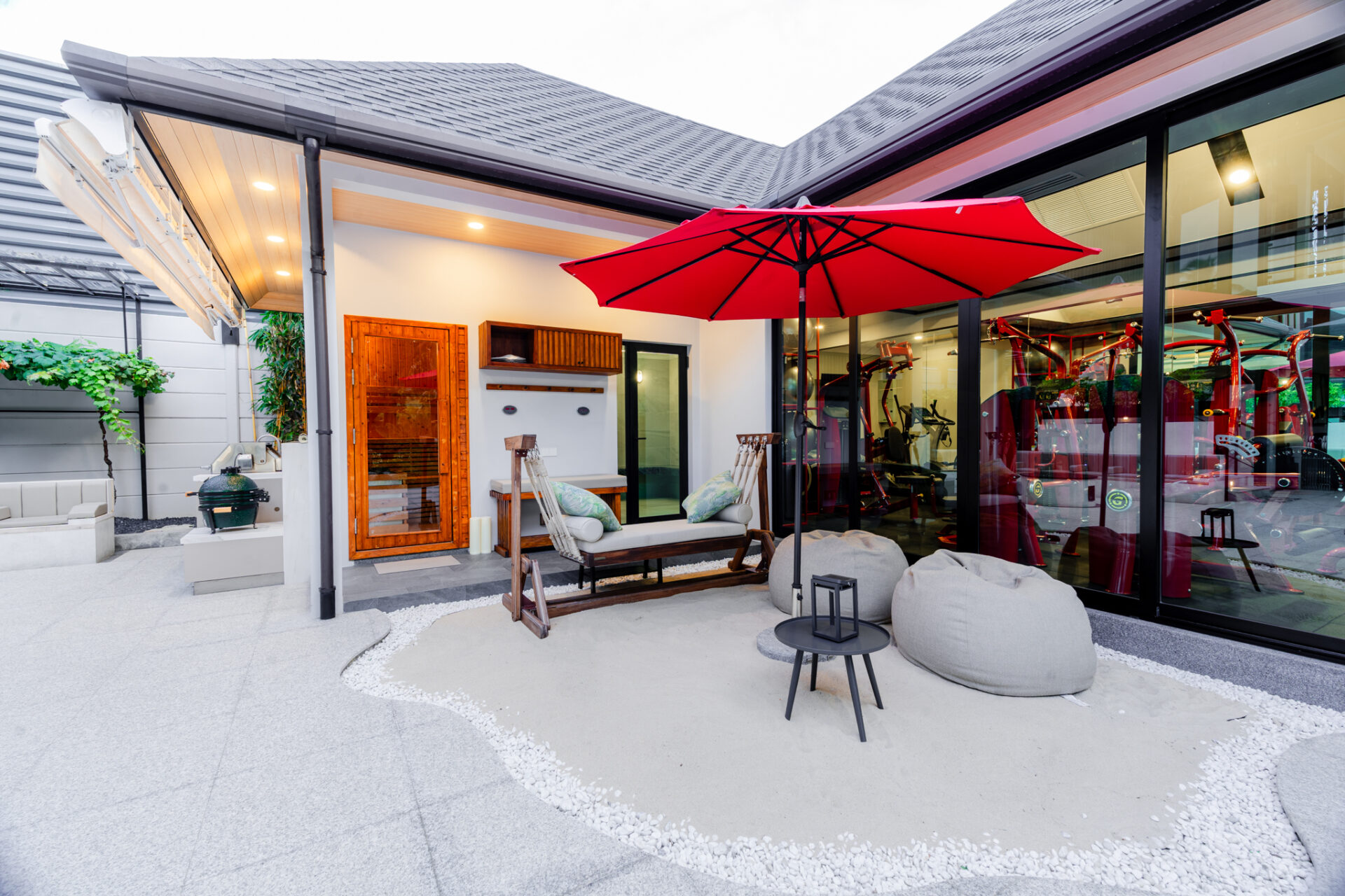Outdoor patio with a red umbrella, wooden seating, and bean bags beside a glass-walled gym area.
