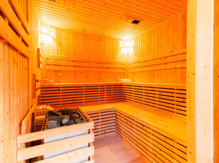 Wood-paneled sauna interior with multiple bench levels and a heater with rocks in the corner, warm amber lighting