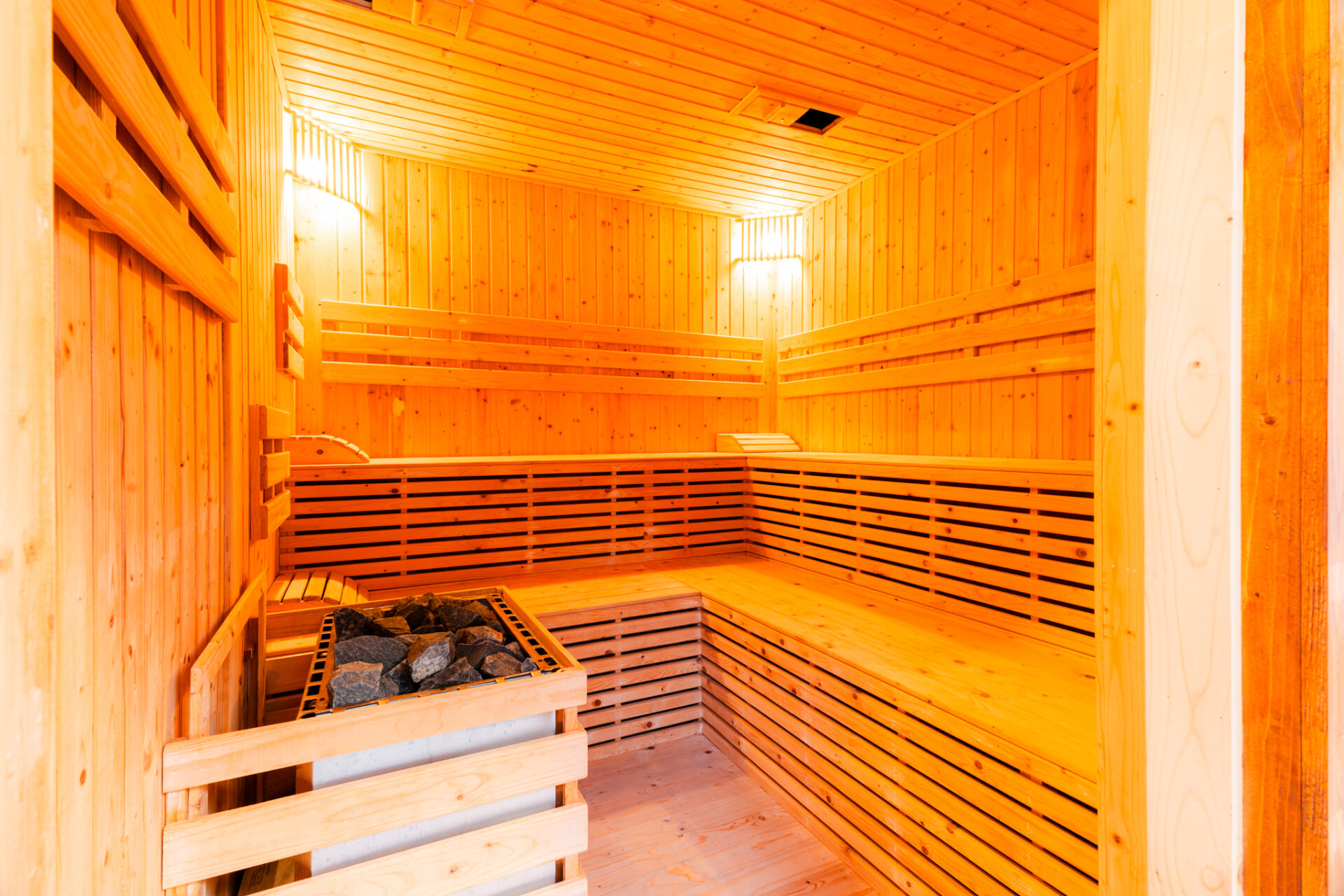 Wood-paneled sauna interior with multiple bench levels and a heater with rocks in the corner, warm amber lighting