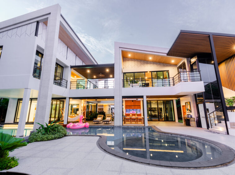 Modern two-story house with a curved pool, glass railings, and outdoor lounge area; a pink flamingo pool float nearby.