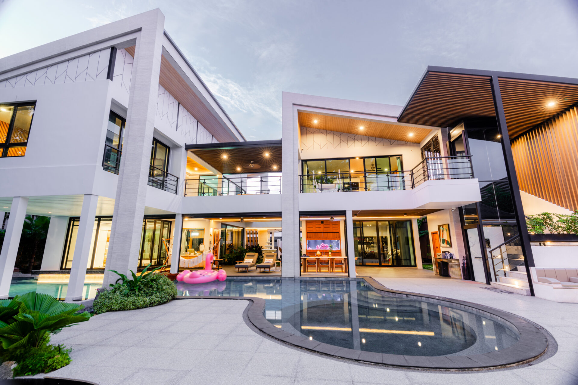 Modern two-story house with a curved pool, glass railings, and outdoor lounge area; a pink flamingo pool float nearby.
