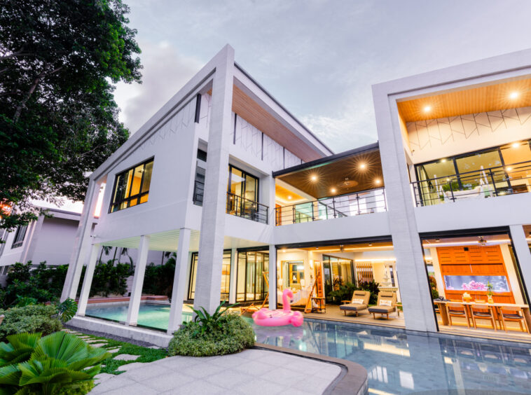 Modern white two-story house with glass walls, pool, and outdoor seating at dusk.