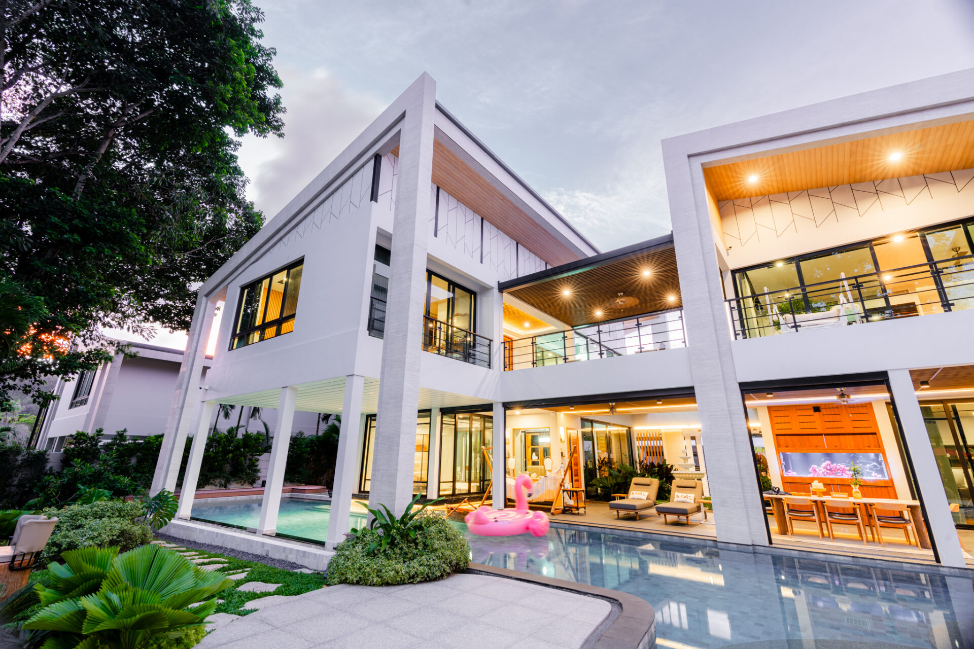 Modern white two-story house with glass walls, pool, and outdoor seating at dusk.