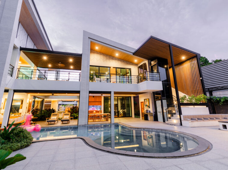 Modern house with glass walls, outdoor seating, and illuminated interiors around a curved pool courtyard at dusk