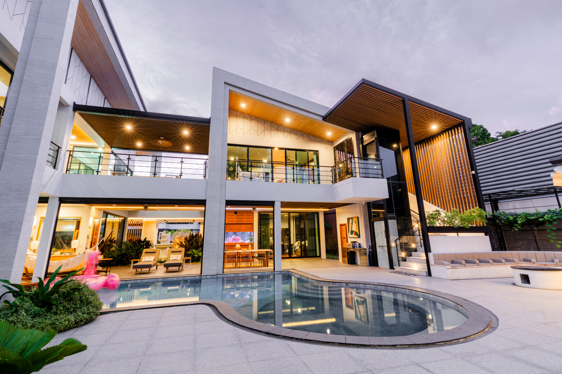 Modern house with glass walls, outdoor seating, and illuminated interiors around a curved pool courtyard at dusk